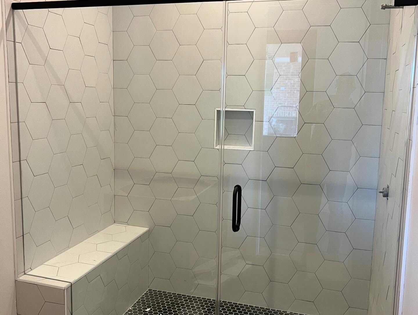 Walk-in shower tiled in large white hexagon tile with a built-in bench, a recessed niche and a black penny round tile shower floor behind glass doors