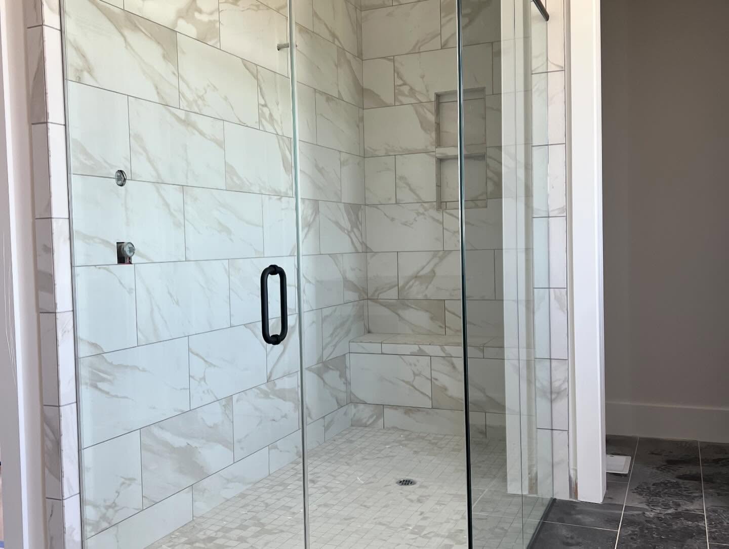 Walk-in shower tiled in large format marble look porcelain with a built-in corner bench, two recessed niches and frameless glass doors with a matte black handle