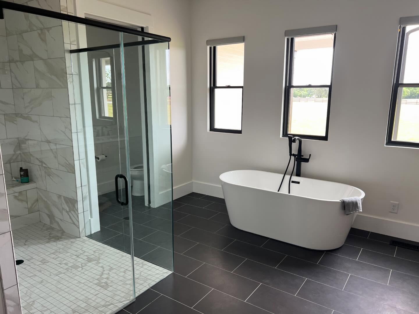 Primary bathroom with a freestanding soaking tub under three windows, matte black filler, and a marble tiled walk-in shower behind frameless glass, over dark slate tile flooring