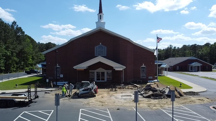 Brick church with a white steeple during construction work to enclose the front covered entrance, with crews and equipment on site