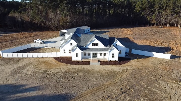 Aerial view of a completed white farmhouse style custom home with a dark roof, fresh landscaping and a fenced yard