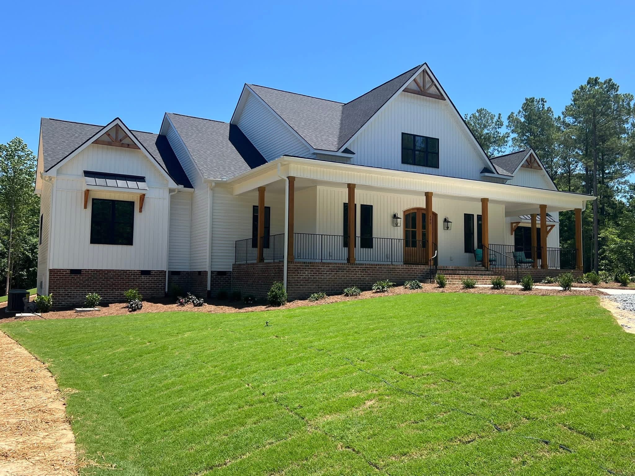 Finished white farmhouse style custom home with cedar porch posts, black windows, metal window awnings, a brick foundation and fresh sod
