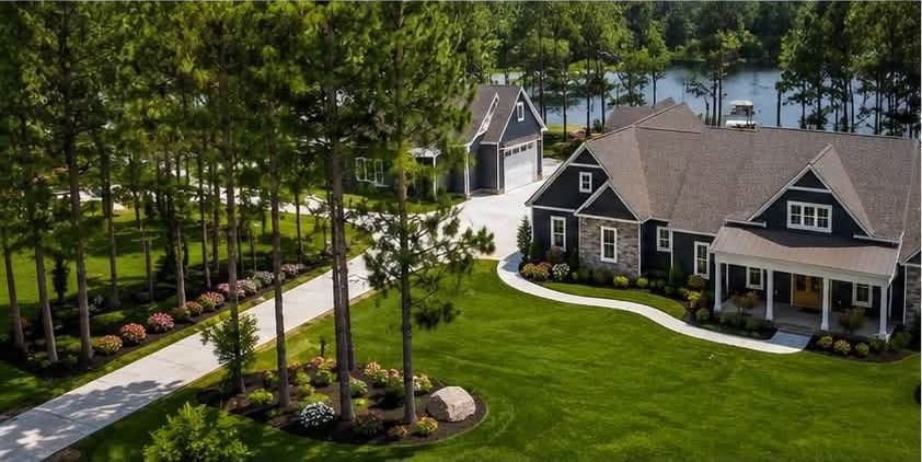 The same navy custom home seen down its concrete drive through pines, with the detached garage and the lake behind