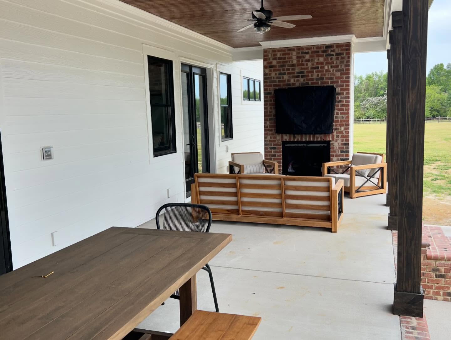 Brick fireplace running floor to ceiling on a covered rear porch, with a mounted television above the firebox, a stained tongue and groove ceiling and seating on the concrete deck