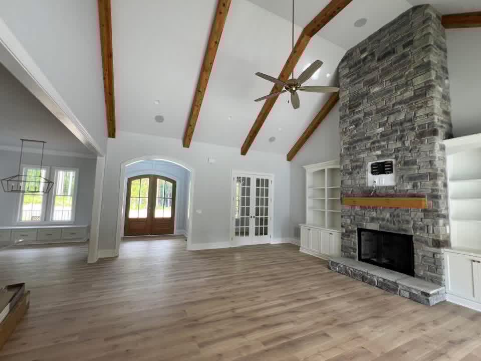 Wide view of the same vaulted great room, showing the full height stone fireplace, cedar beams, French doors and the arched entry to the foyer