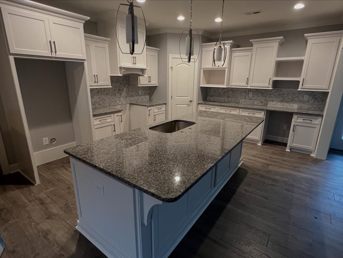 Finished custom kitchen with a large granite island with an undermount sink, white cabinetry to the ceiling, herringbone tile backsplash and black pendant lights