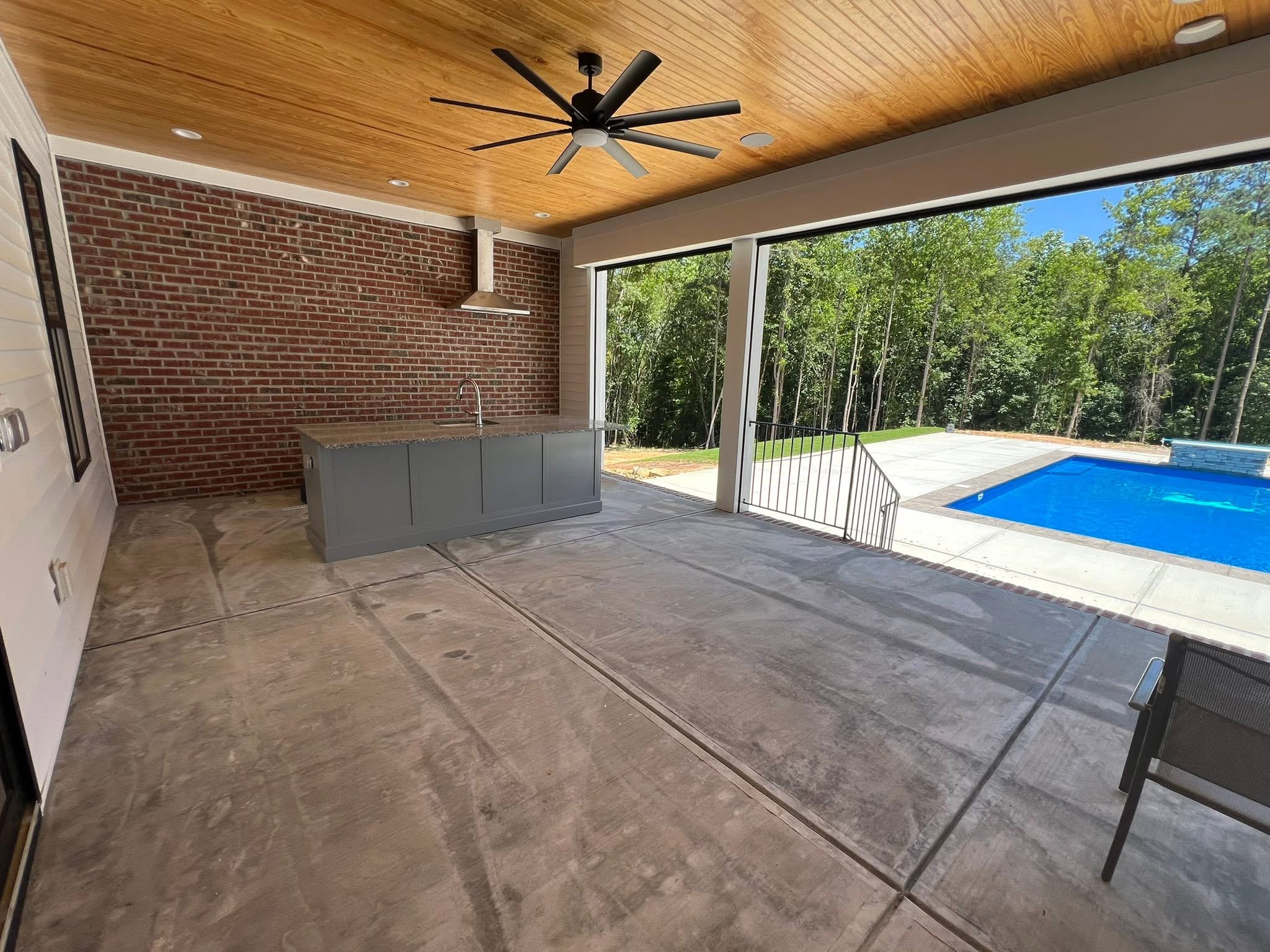 Covered patio with a stained tongue and groove ceiling, brick wall, outdoor kitchen island with a sink and vent hood, and an in-ground pool beyond