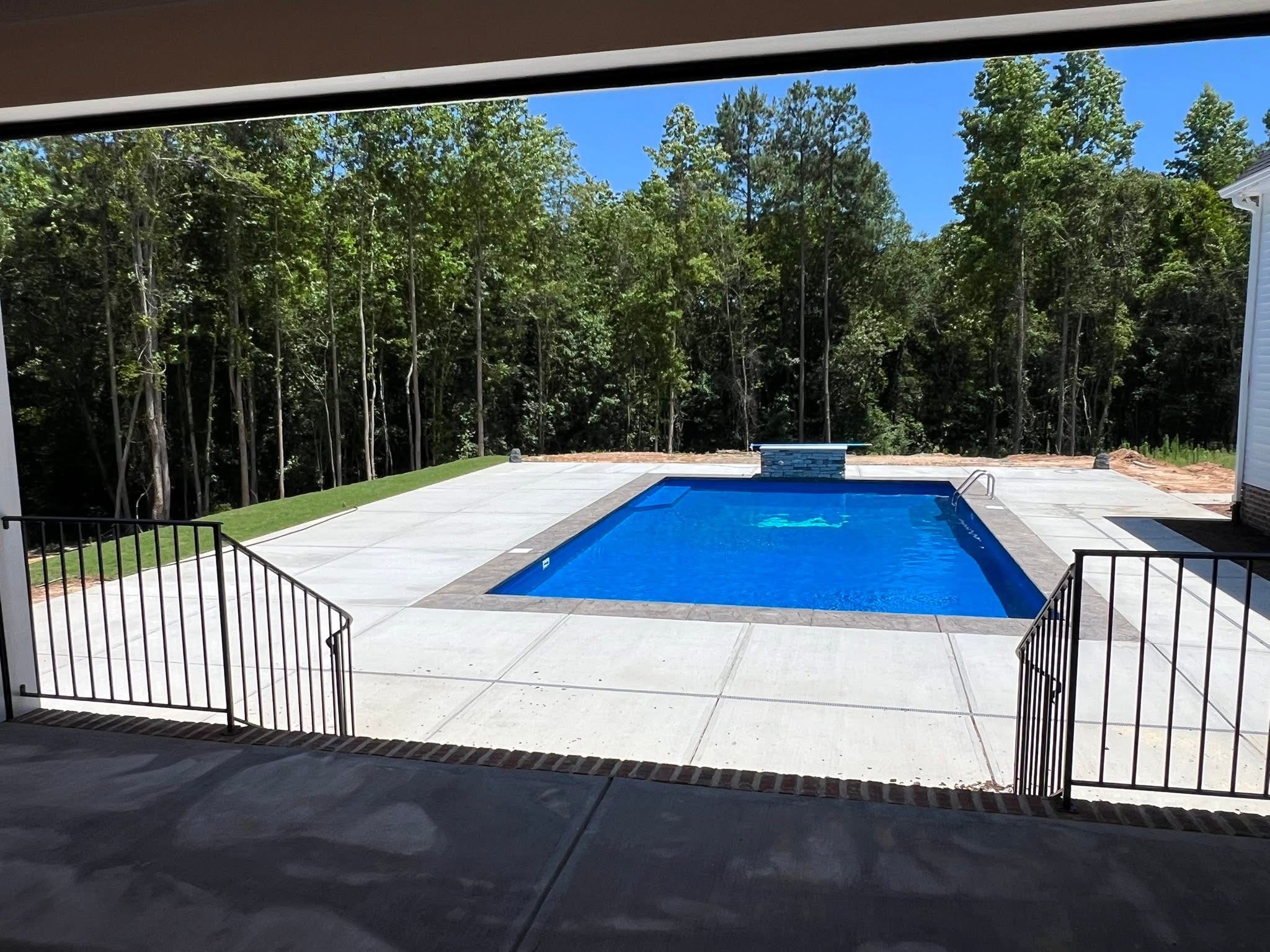 In-ground pool with a diving board and stone water feature, surrounded by a poured concrete deck with a stamped border, seen from a covered porch