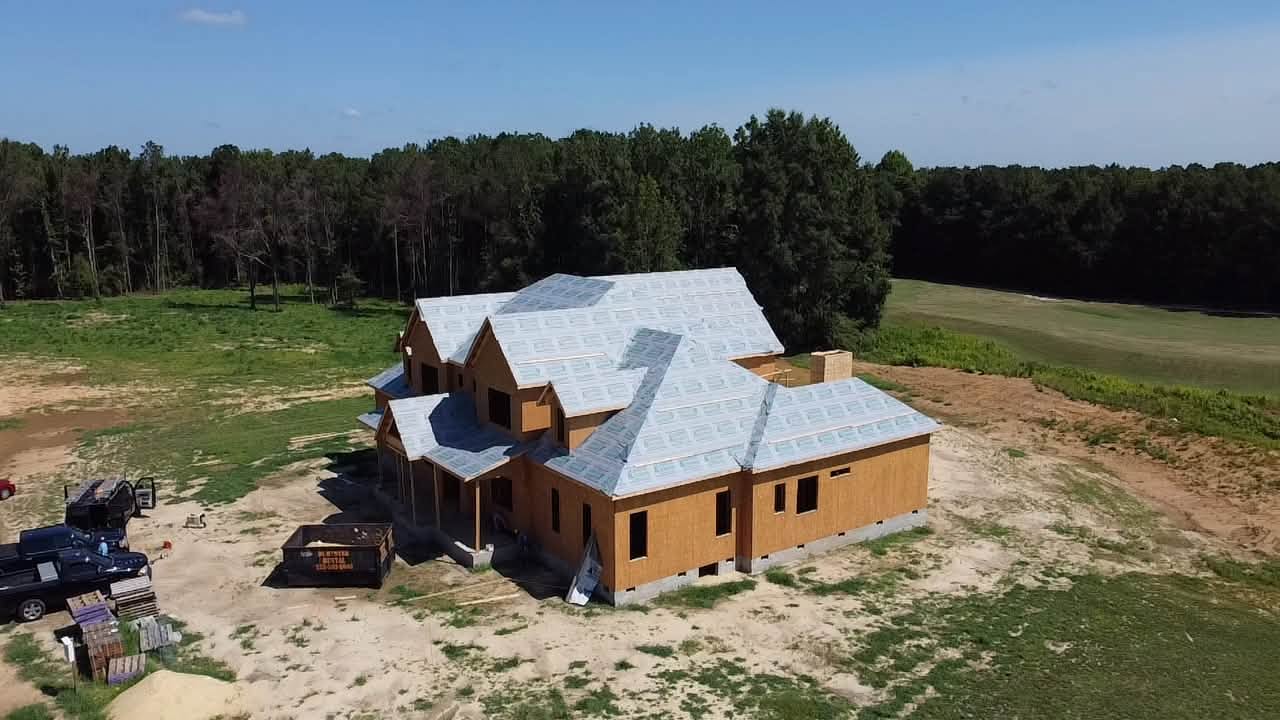 Aerial view of a single story custom home under construction on a cleared lot bordered by pine woods